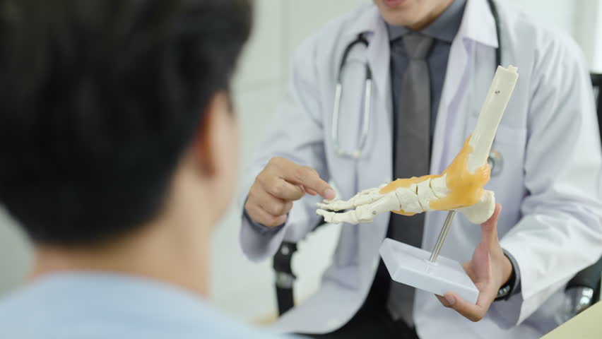 Doctor explaining foot joint anatomy to patient using realistic bone model during orthopedic consultation, healthcare professional demonstrating bone structure and discussing foot or ankle condition.