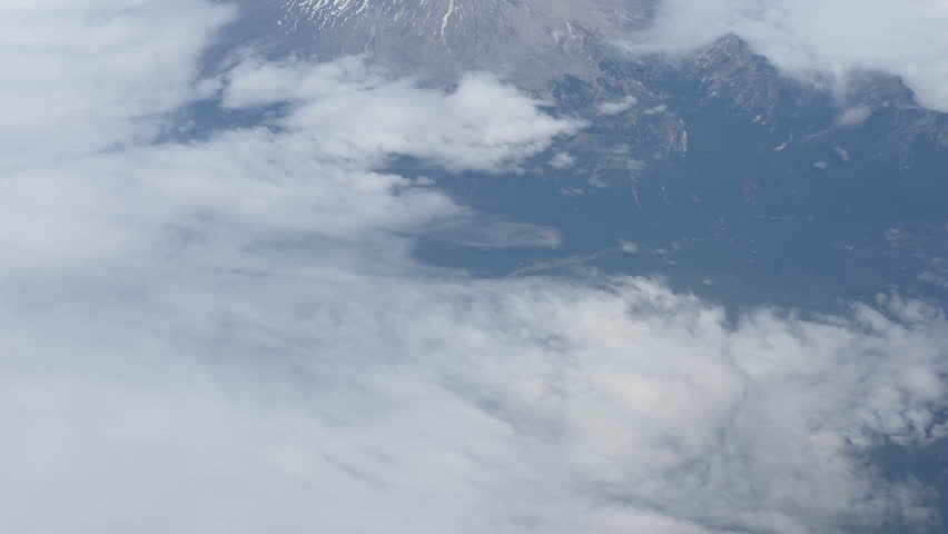A tilting up aerial view of a partially snow-capped Mount St. Helens, Washington, surrounded by broken cloud layers