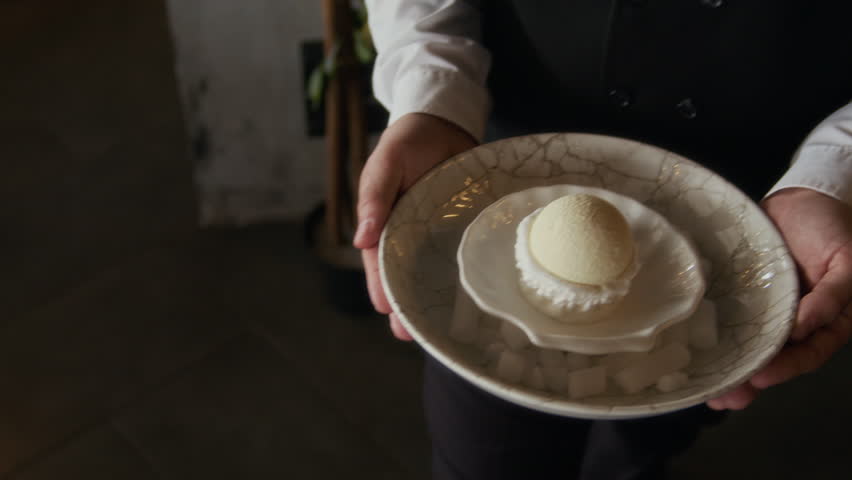Cropped shot of hands of unrecognizable waiter in formal attire presenting sophisticated white dome dessert served on shell-shaped plate to guests drinking wine in restaurant