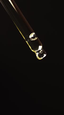 Vertical macro shot of yellow oil or serum drop falling from pipette with air movement. Slow motion product footage on black background for premium skincare use.