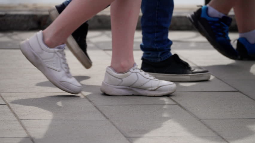 Close-up sequence showing a group of people walking on an outdoor paved walkway, focusing on their legs and various sneakers in motion during daylight.
