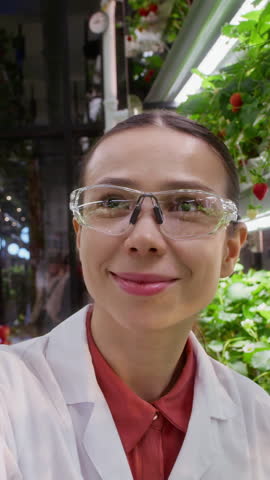 Vertical shot of young smiling female farmer wearing protective eyeglasses looking at you while making livestream from vertical farm and talking about harvest of new sorts of strawberry