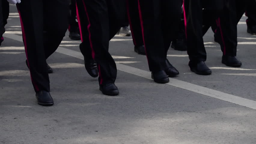 Close-up view of a group marching in unison, focusing on synchronized footsteps and formal uniforms with red stripes on an urban street setting