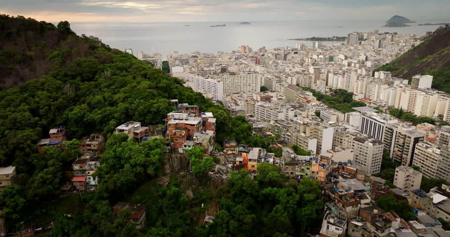 Aerial drone view of social inequality in Rio de Janeiro, dense favela on lush green hill contrasts with modern buildings of Copacabana. Brazil