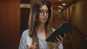 Young hispanic woman wearing glasses and striped shirt holds a creditcard and taps a tablet in a hotel corridor; convenience. - Powered by Shutterstock - Get 15% off with code: PIKWIZARD15