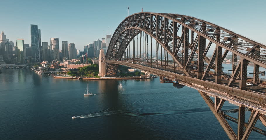 Australia, Sydney: Sydney Harbour Bridge and city skyline, sailboat glides under sunny morning blue sky. Travel destination, modern cityscape architecture in background. Aerial view drone footage