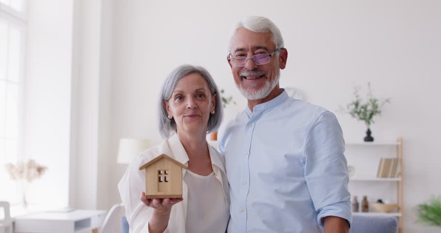Happy senior smiling couple moving new flat, holding toy house showing keys from new own house, getting bank loan for older citizen, elder family making deal with property real estate purchase dream