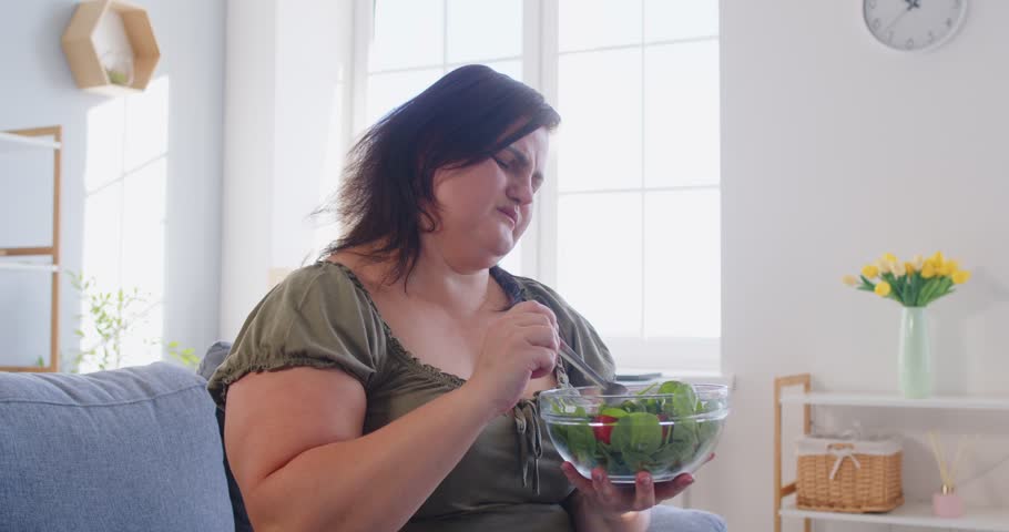 Young fat overweight sad woman tired of diet restrictions eating healthy salad with unhappy face expression sitting on sofa at home. Nutrition, dieting and weight loss concept. 4k video.