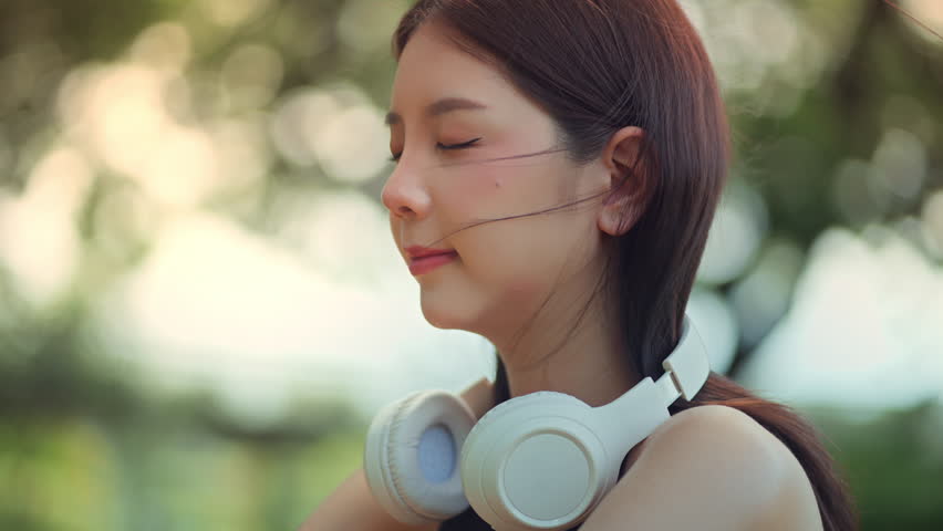Close-up of peaceful asian woman with headphones looking up calmly while enjoying gentle breeze in city park – wellness lifestyle, good health and positive mental wellbeing in relaxing urban nature
