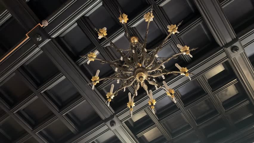 A grand, ornate golden chandelier with multiple candle-like lights hangs from a dark, coffered ceiling, showcasing intricate historical design and creating a rich ambiance