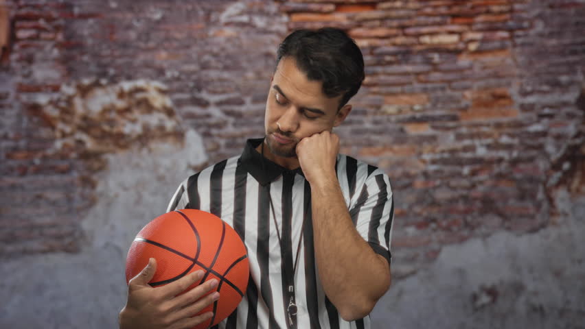 Young hispanic man in referee uniform holding basketball pensively against isolated brick background, reflecting thoughtful sporting concept.