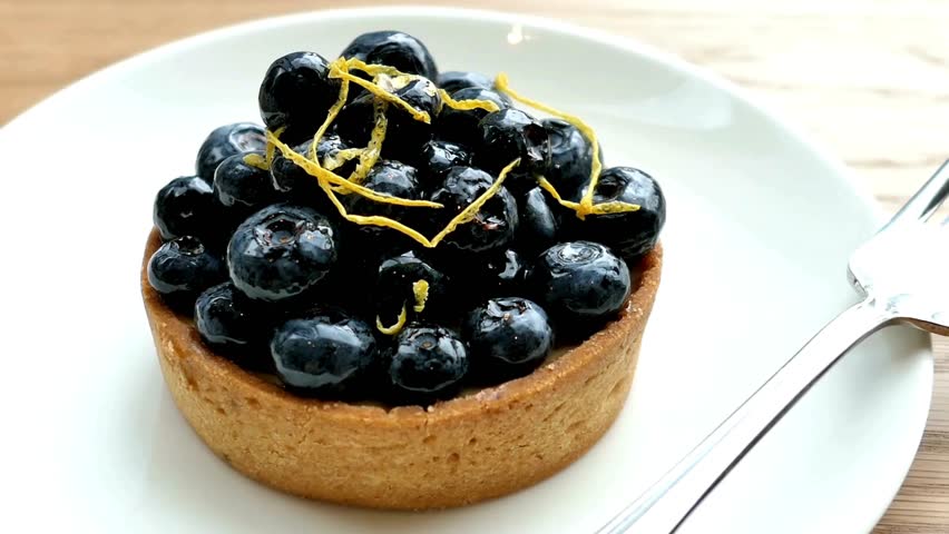 A blueberry tart topped with lemon zest sits on a white plate