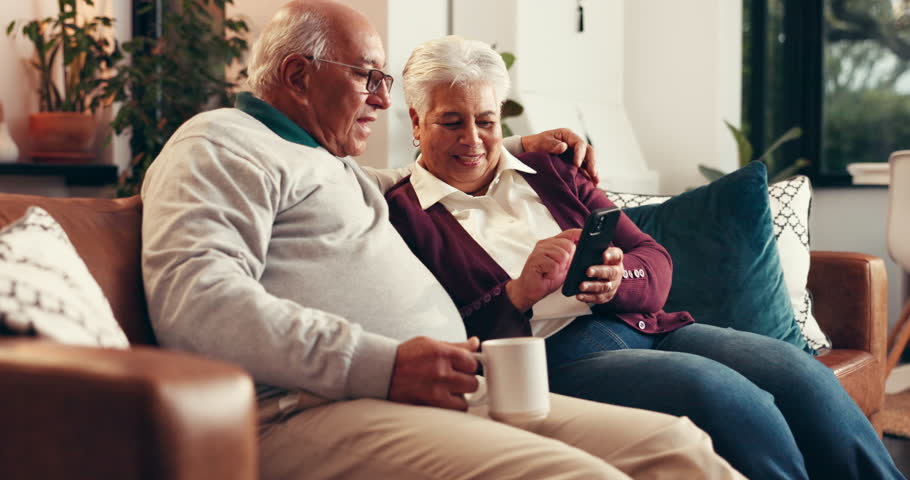 Coffee, mobile and elderly couple in home for relax, conversation and happy together with embrace. Drink, man and woman in retirement with phone for reading news, article and puzzle app on internet