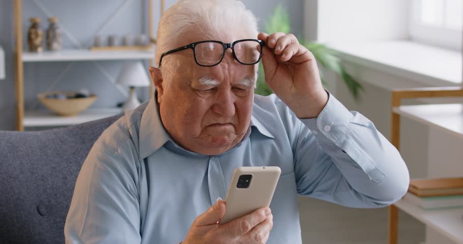 Elderly man has bad eyesight, puts off eyeglasses and focused in smartphone screen, tries to read received message, cannot understand how to use app, has some problems. Technology annd retirement