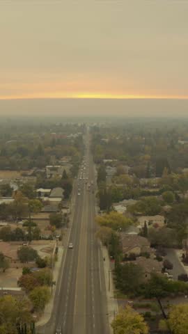 Aerial view of Carmichael, California at sunset. A straight road stretches through a suburban neighborhood surrounded by autumn-colored trees under a hazy sky (Vertical 4K for Reels, Tiktok, Shorts)