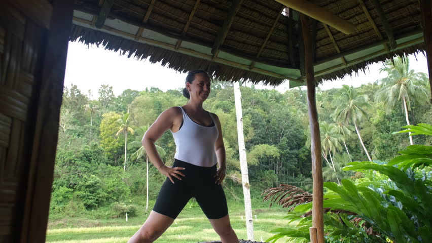 Young woman performing yoga poses on a bamboo hut terrace, surrounded by scenic rice fields and vibrant tropical plants, slow motion. Active lifestyle and wellness concept