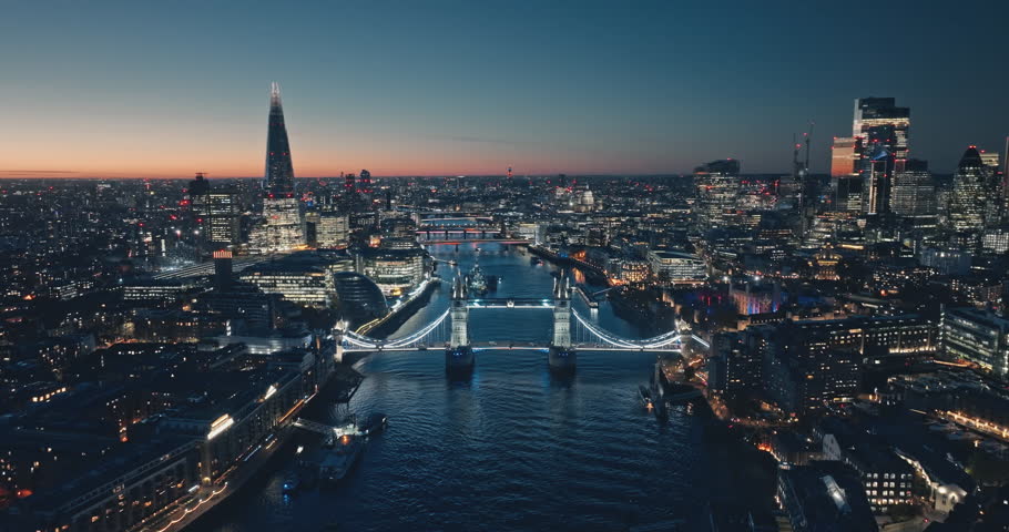 London: Aerial view of city illuminated skyline at sunset, iconic Tower Bridge, The Shard, and night cityscape stretching along the River Thames. Drone flight footage zoom out panorama. Blue toning