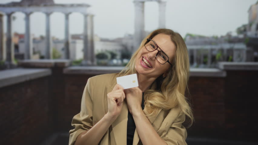 Woman smiling outdoors holding credit card with ruins in background, wearing glasses and blazer, showcasing travel excitement in ancient european setting.