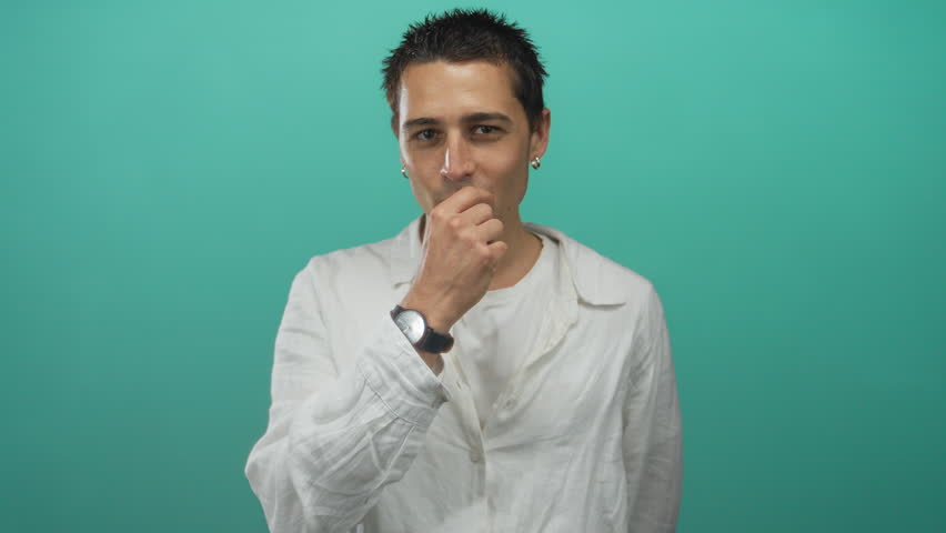 Young hispanic man making zip lips gesture in studio against green wall, wearing white long sleeve shirt and wristwatch; silence.