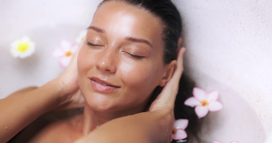 Slow motion close-up of a tanned woman gently caressing her face in a luxury spa bath with tropical flowers. Ideal for skincare, wellness, treatment, and beauty product promos.