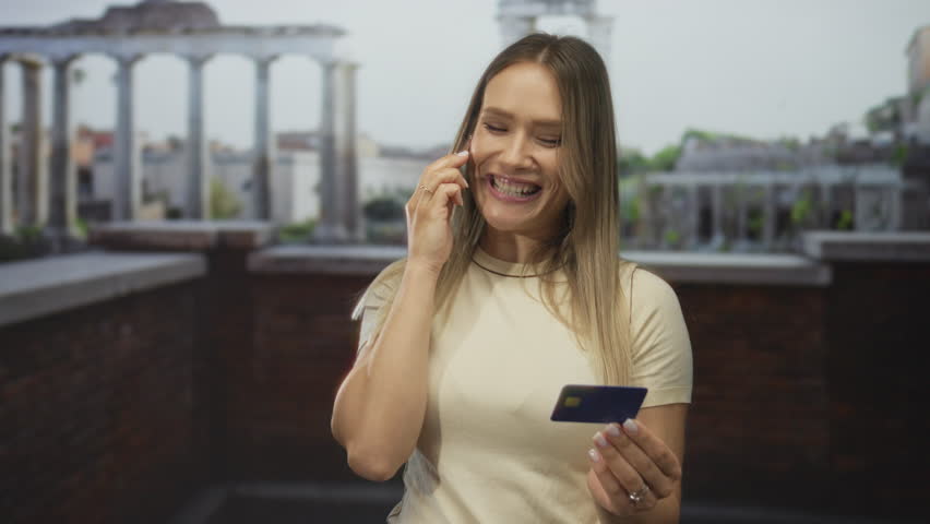 Woman smiling while talking on smartphone and holding credit card against backdrop of ancient roman ruins in city setting under outdoor light