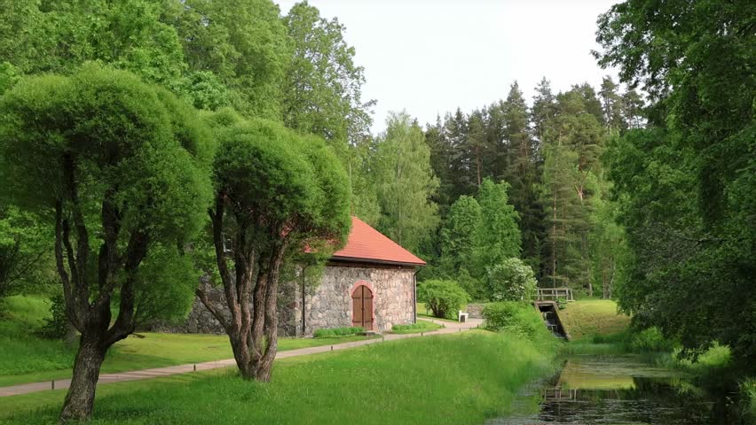 Charming stone cottage with red roof stands by calm stream in lush green forest, surrounded by manicured grass and shaped trees. Peaceful countryside retreat. Picturesque landscape with a rustic house