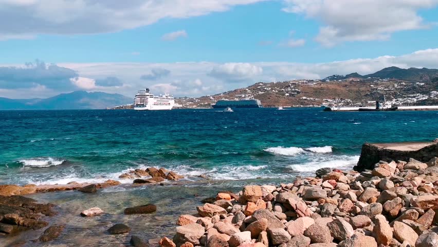 View of rocky beach in Santorini, Greece, with turquoise sea waves hitting shore, cruise ships in distance, and scenic coastal hills beneath a vibrant blue sky with scattered clouds