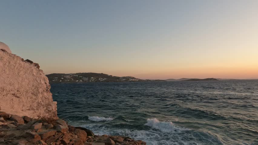 Panning shot of sunset over sea in Santorini, Greece, with waves crashing near white coastal chapel wall, blue window shutter, and cross silhouetted against warm evening horizon