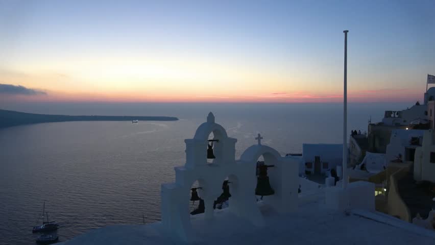 A breathtaking aerial view captures the serene sunset over Santorini, Greece. The iconic white bell towers stand against the vibrant horizon, creating a tranquil, picturesque scene.