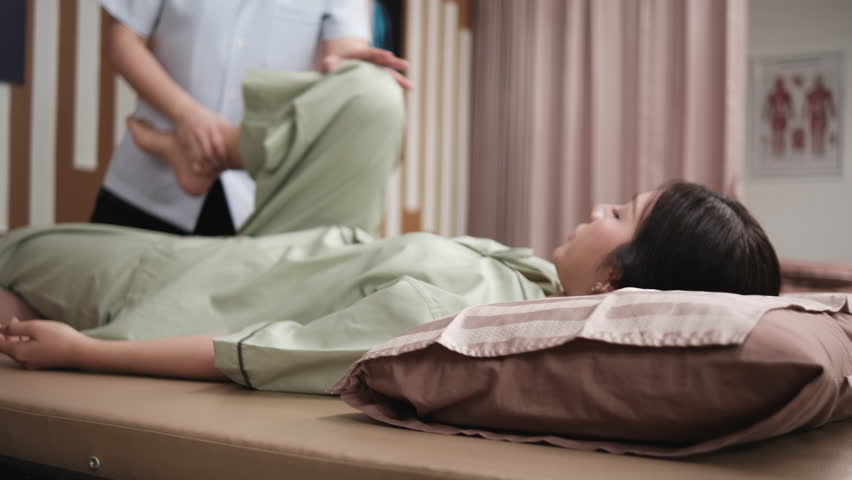 Asian female patient's legs were stretched on massaging bed, treated for health recovery, knee muscle pain relief by physical therapist, and passive medical therapy at healthcare wellness spa clinic.