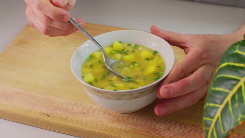 Pea soup with potatoes and chopped green onions is stirred and brought to mouth with a spoon. Close-up of homemade soup tasting in a cozy kitchen setting.