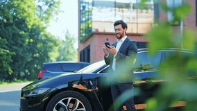 Successful businessman in smart suit charges sleek black electric vehicle outdoors. Engaged with smartphone, displaying satisfaction and embracing modern, eco-friendly transportation solutions. - Powered by Shutterstock - Get 15% off with code: PIKWIZARD15