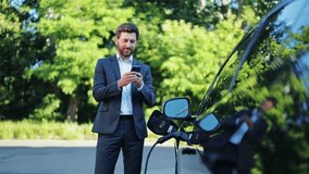 Professional businessman in dark suit confidently checks smartphone as modern electric vehicle charges outdoors. Focused and content, reflecting smart, eco-friendly lifestyle. - Powered by Shutterstock - Get 15% off with code: PIKWIZARD15