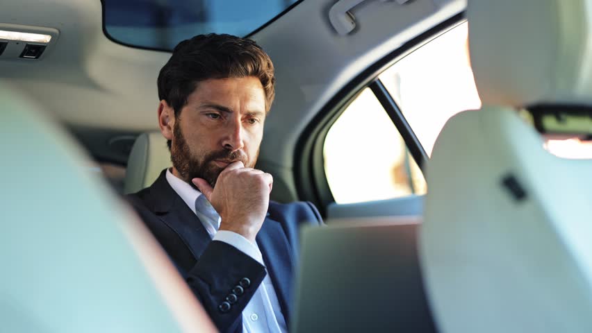 Professional businessman, dressed in a sleek suit and white shirt, thoughtfully uses his laptop in a luxurious car backseat. His expression shifts from focused concentration to satisfied success.