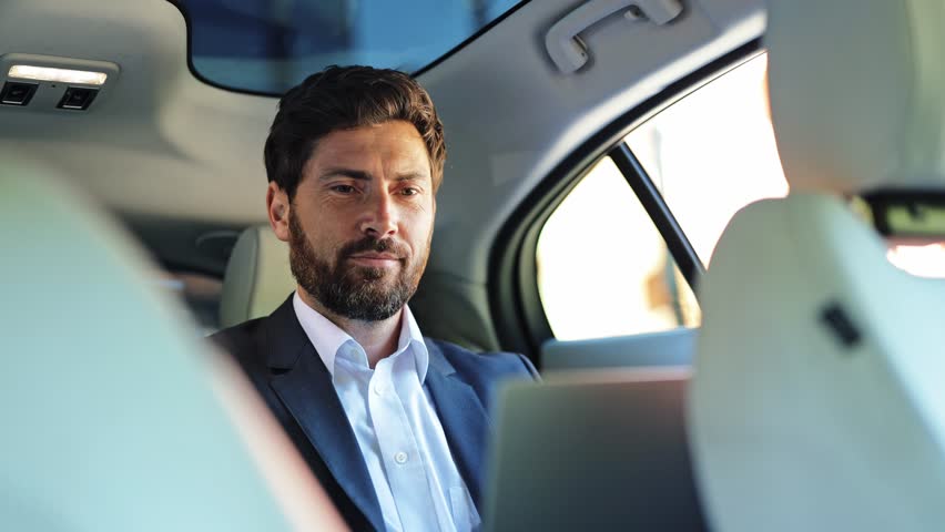 Professional businessman, dressed in a sleek suit and white shirt, thoughtfully uses his laptop in a luxurious car backseat. His expression shifts from focused concentration to satisfied success.