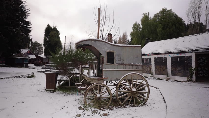 Static camera shots antique wooden horse-drawn carriage covered in snow at Colonia Suiza, Bariloche with traditional Andean setting and wintry landscape evoke rustic heritage charm during snowfall.