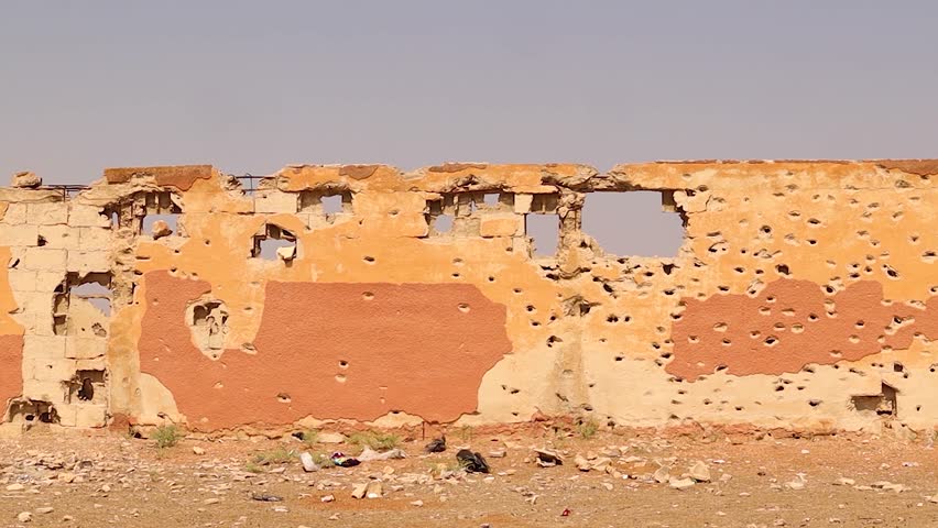 A shattered, bullet-riddled wall stands across from Aleppo Central Prison, bearing scars of the fierce Syrian war. A powerful visual of destruction, resilience, and post-war urban history.