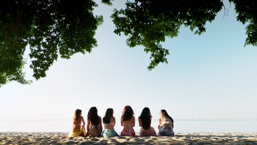 A group of girls sit on the sand near the water, chatting and raising their hands up against the backdrop of a calm sea at sunset. Women enjoy communication and a beautiful view of nature