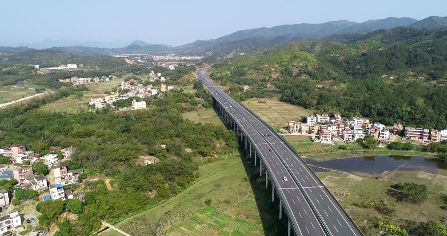 This aerial perspective showcases a modern highway winding through a serene countryside in southern China. Rolling hills and a peaceful village are visible, offering a glimpse of rural life.