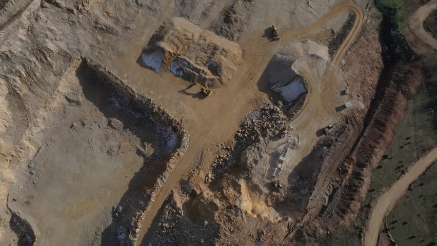 Top-down aerial view of a large open-pit quarry with heavy machinery. Excavators and loaders process stone, creating sand and gravel stockpiles for construction.
