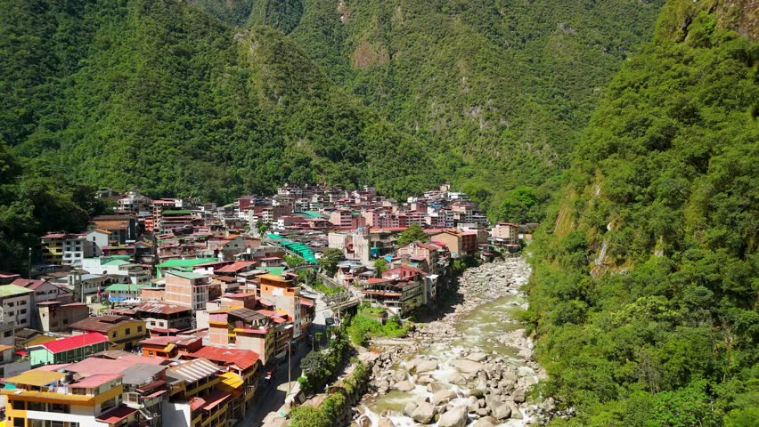 Aerial pullback of Aguas Calientes town nestled in dense green forest at the base of Andean peaks