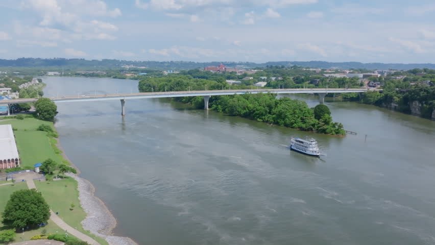 An aerial view of the Tennessee River in Chattanooga, TN, shows a riverboat cruising near a bridge and a lush island under a partly cloudy sky.