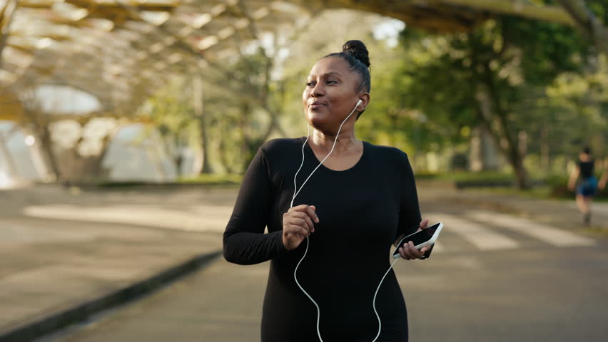 Happy Adult Girl Jogging Outside in the Park with Earphones in Her Ears and Listening to Music While Training. Positive Jogger Burning Calories Outdoors Doing Cardio. People and Sport Concept