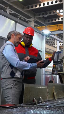 Production Manager Discussing about Job with Black CNC Machine Operator in a Factory. Middle-Aged Manager with Gray Hair, Using Digital Tablet in Communication with Young Worker. Industrial Leadership