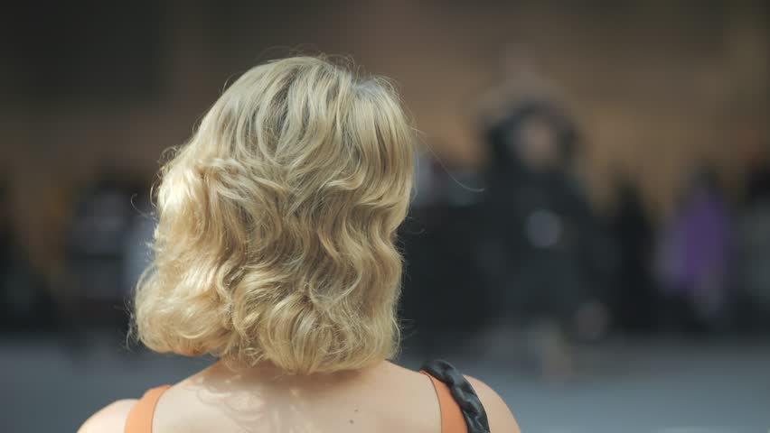 Blonde woman with a short haircut is looking at the camera. She is wearing an orange top and a black purse. The scene appears to be a casual gathering or event