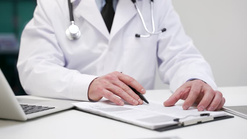 Close up. Male doctor signing medical documents in hospital office. Physician in lab coat reviewing a report on clipboard, writing prescription and filling out patient records or treatment conclusion