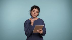 Thoughtful curly young woman notes in paper notebook, future plans and to-do list. Pensive beautiful creative female writing creative idea, looks at camera with smile on isolated light blue background - Powered by Shutterstock - Get 15% off with code: PIKWIZARD15