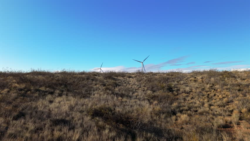 Wind turbines spread across wide industrial and rural grassland under clear vibrant blue sky, Patagonia, Argentina.
