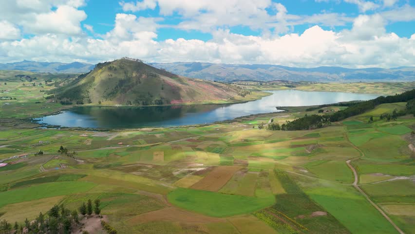 Tranquil landscape of the Peruvian Andes, showing a serene mountain lake, green farm plots, and cloud shadows moving across the land on a sunny day.