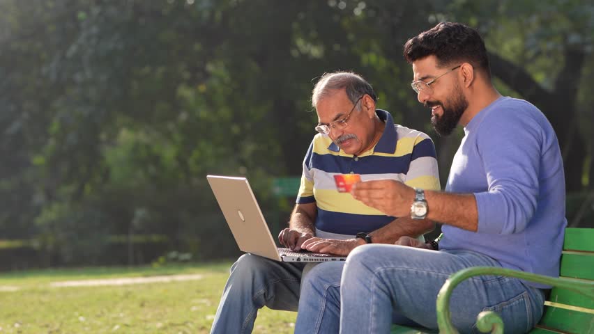 Young indian man showing bank card and laptop how to uses to his father at park